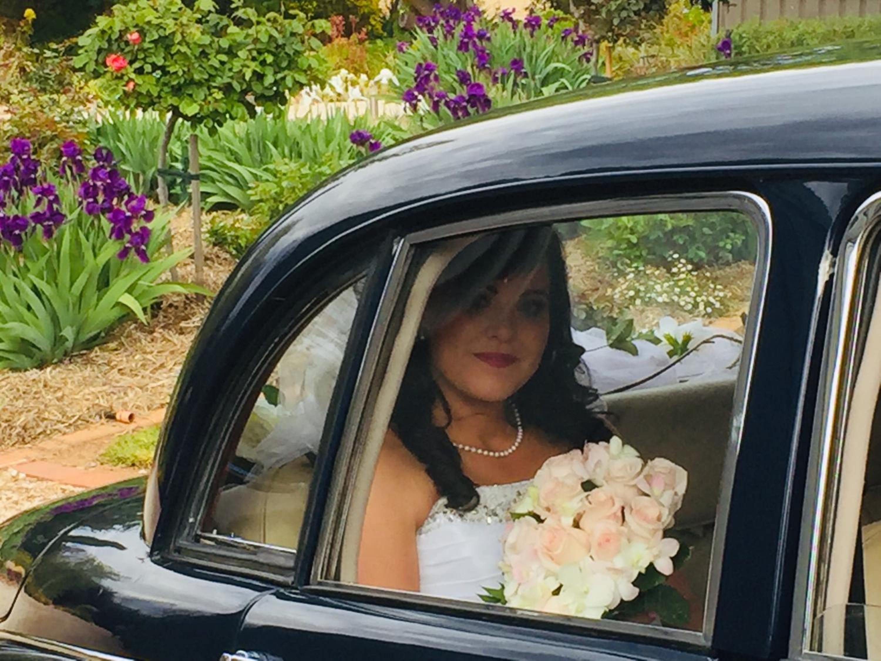 Un coche antiguo con una mujer en vestido de novia sosteniendo un ramo, estacionado en un jardín lleno de flores.