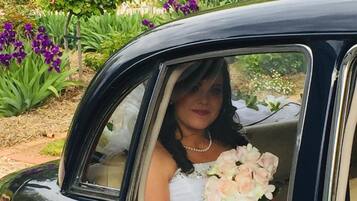 Un coche antiguo con una mujer en vestido de novia sosteniendo un ramo, estacionado en un jardín lleno de flores.