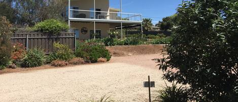 Una casa de dos pisos con balcón, rodeada por un jardín con diversas plantas y un camino de entrada de grava.
