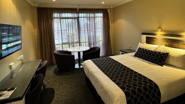 Ein Hotelzimmer mit Bett, Schreibtisch mit Computer, Stuhl und einem Fenster mit Vorhängen.