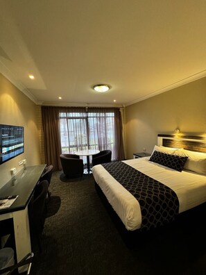 Quarto de hotel com uma cama, uma escrivaninha com computador, uma cadeira e uma janela com cortinas.