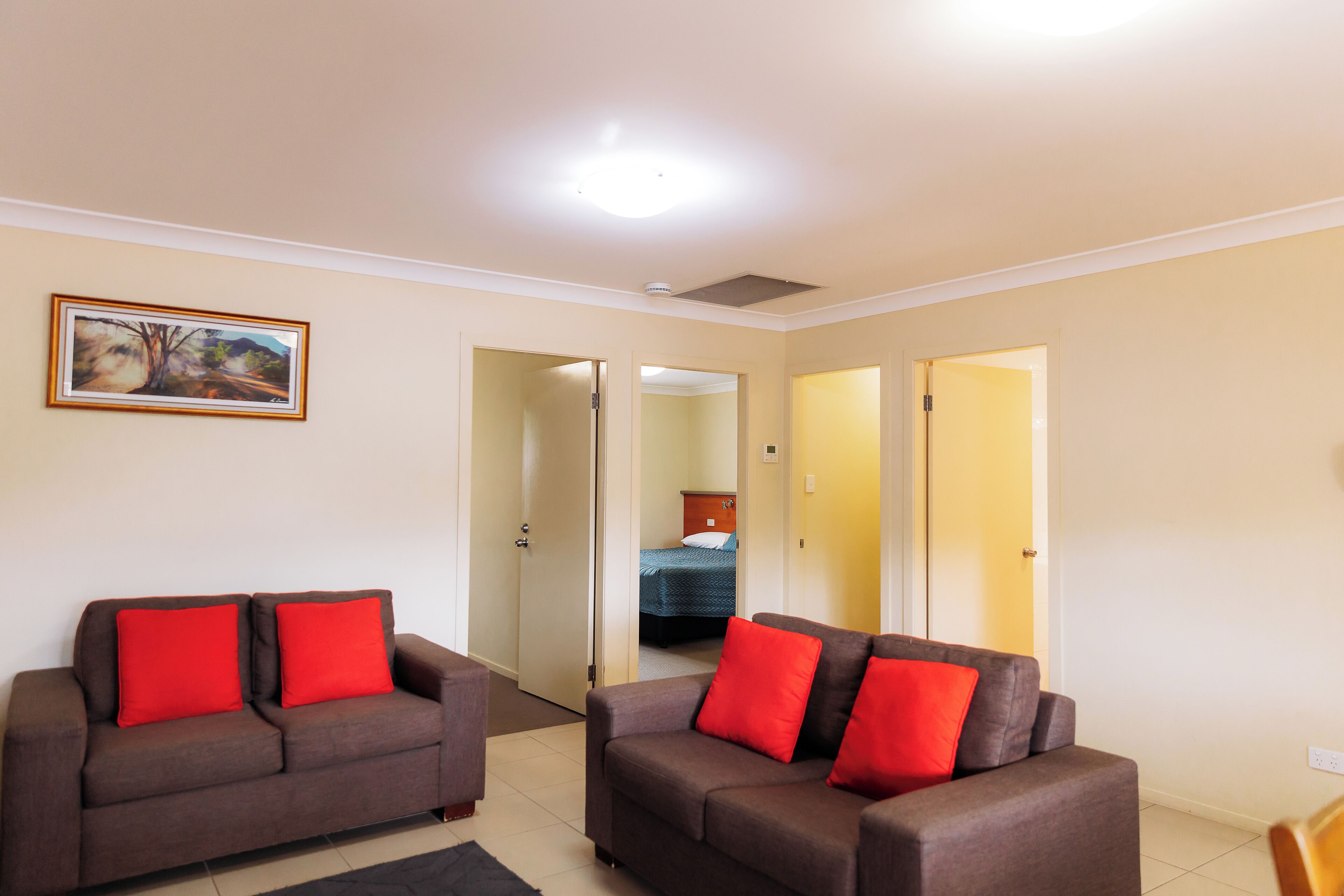 A modern living room with two sofas and red cushions, a painting on the wall, and a bedroom visible through an open door.