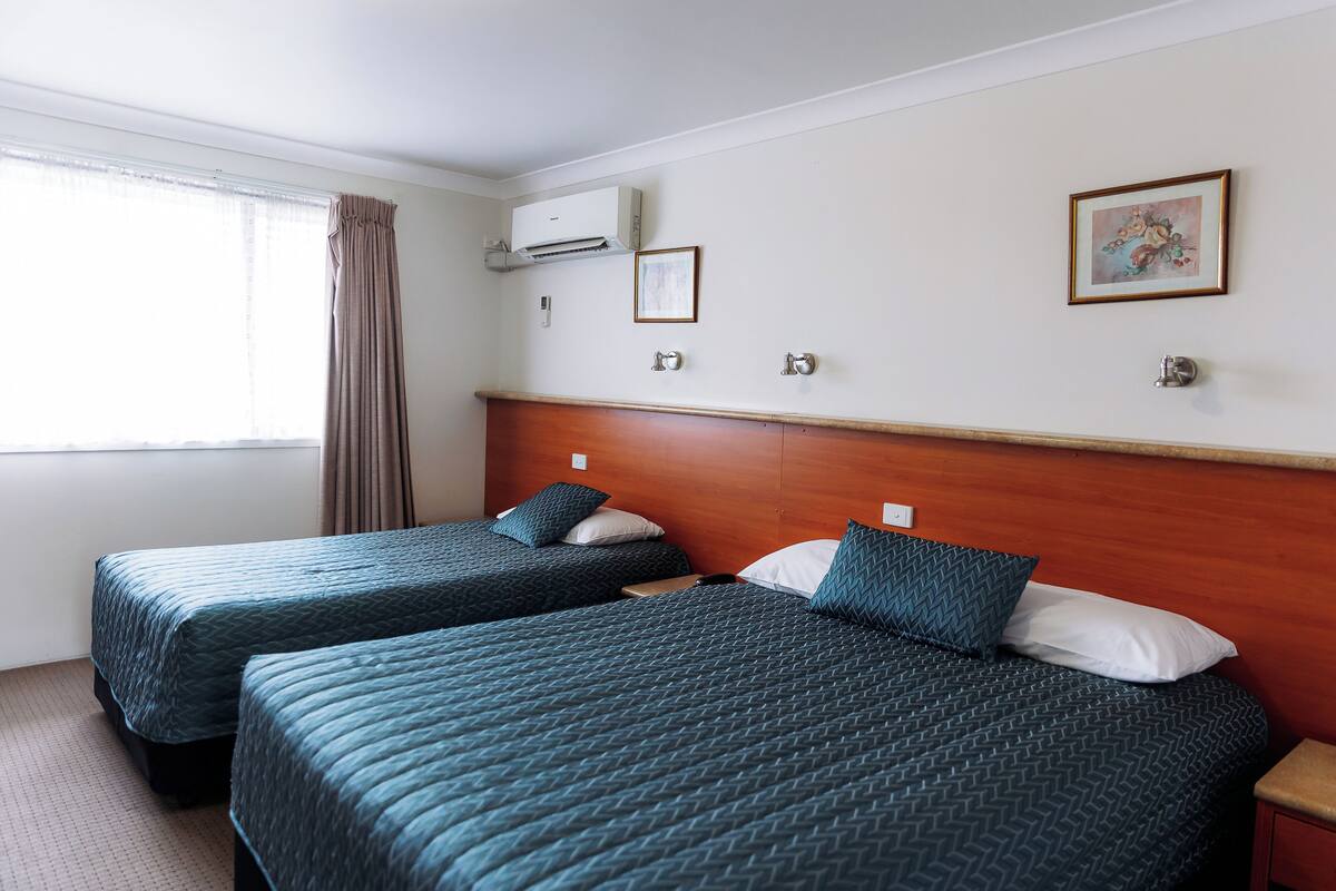 A hotel room with two beds, a wooden headboard, a window with curtains, an air conditioning unit, and a framed picture on the wall.