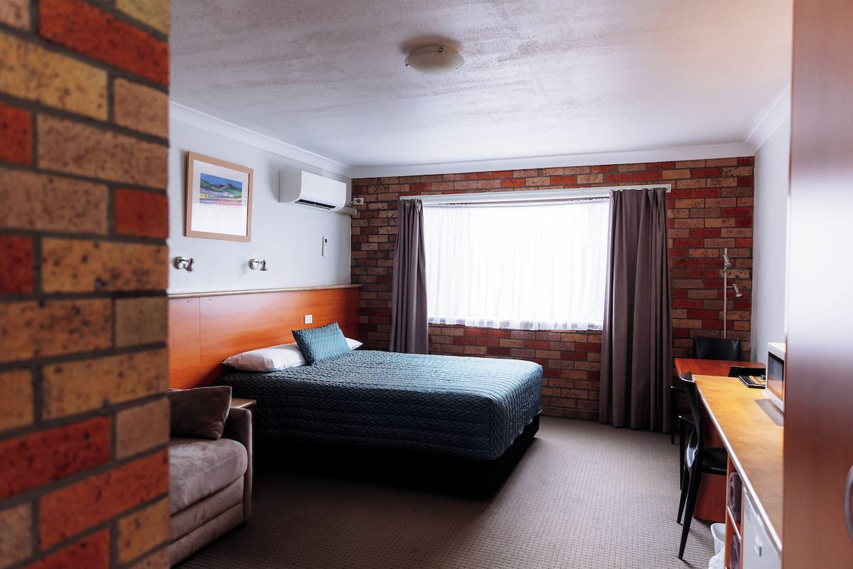 A hotel room with a brick wall, a bed, a desk, and a window with curtains.