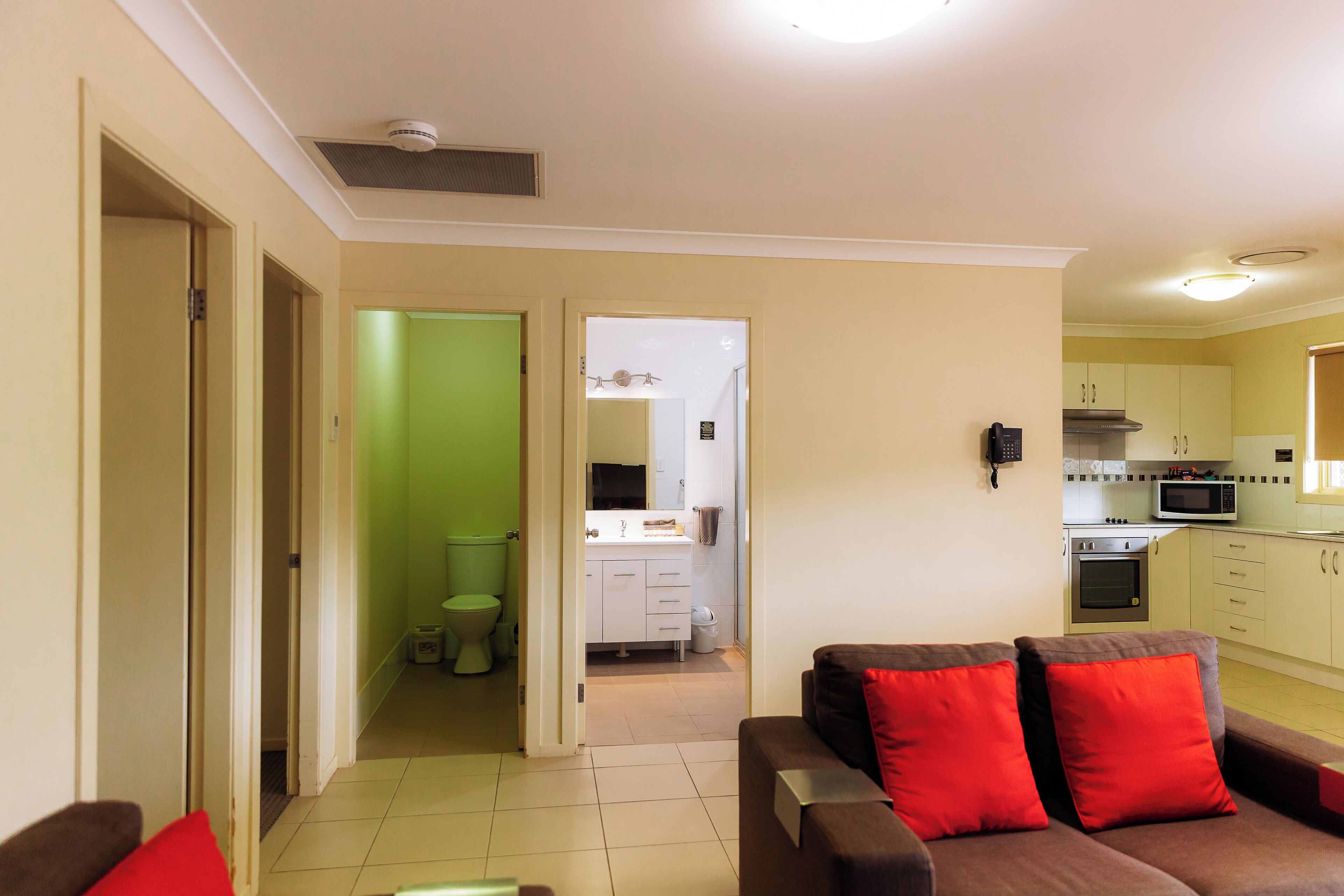A living room with a brown sofa and red cushions, a kitchen with white cabinets and appliances, and a green bathroom.