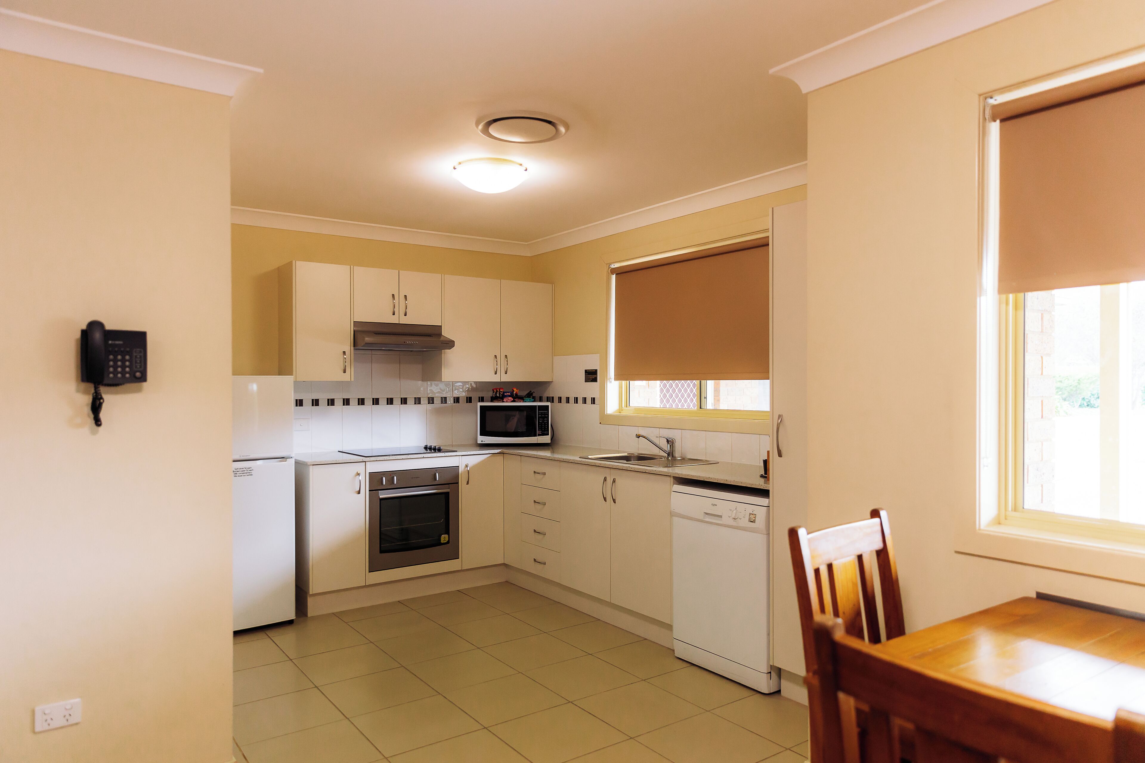 A kitchen with white cabinets, a microwave, an oven, and a dishwasher. There is a telephone on the wall and a window with a blind.