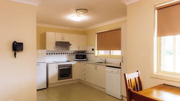 A kitchen with white cabinets, a microwave, an oven, and a dishwasher. There is a telephone on the wall and a window with a blind.