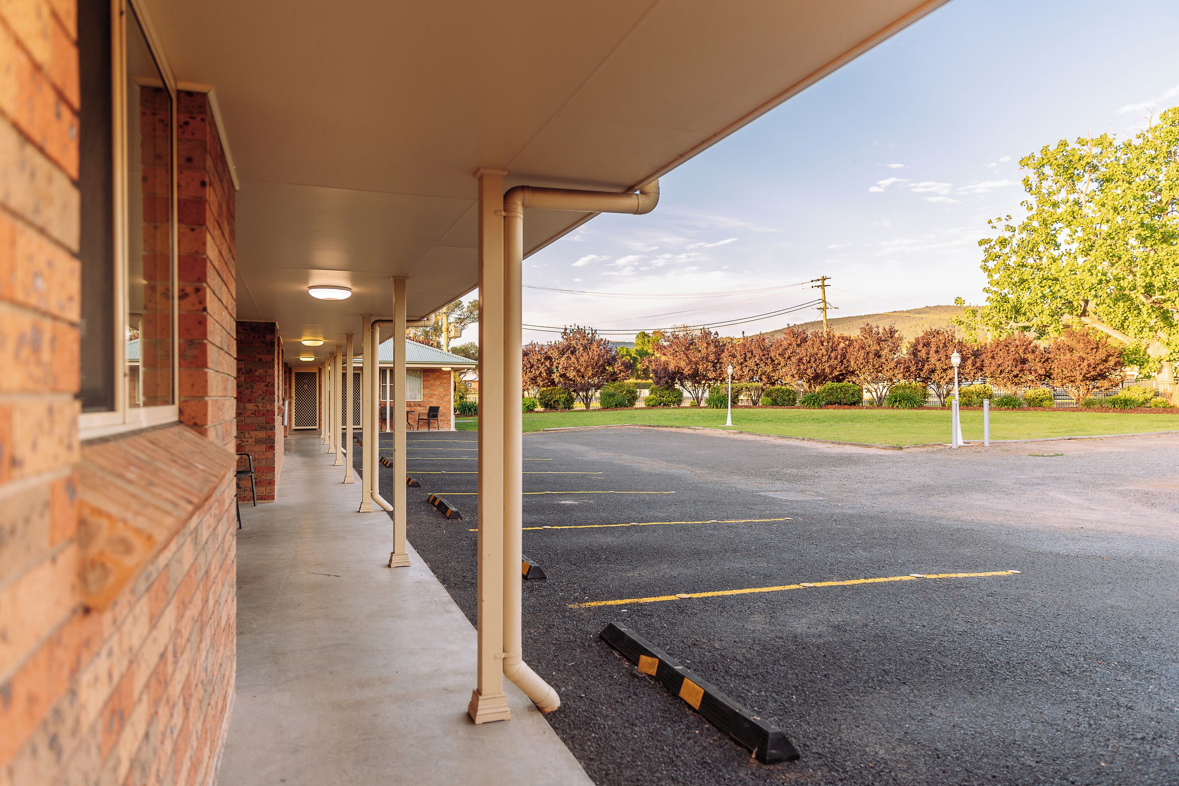 A covered outdoor area with a brick building, a paved parking lot, and a view of a green landscape with trees and a hill.
