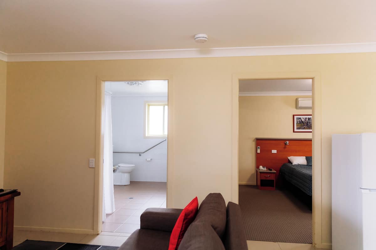 A hotel room with a brown sofa, a bed, a small bedside table, and a white refrigerator.