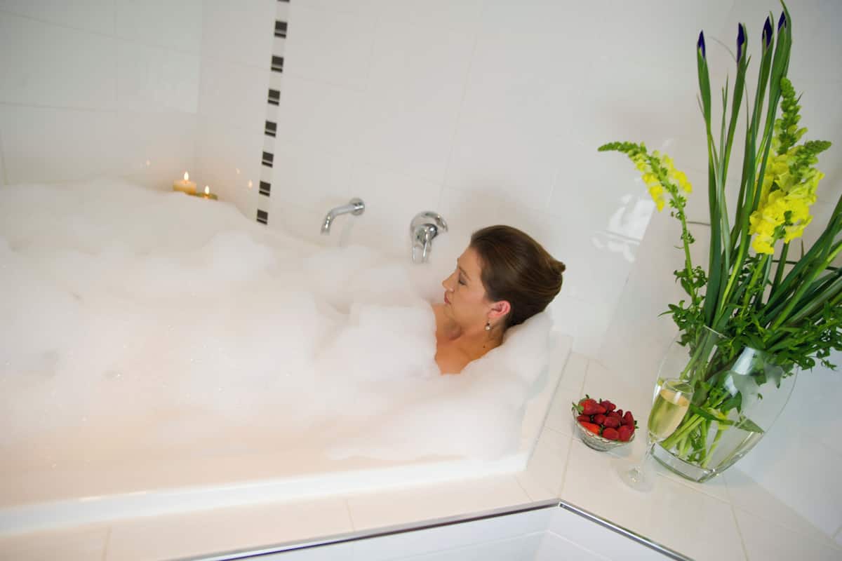 A woman in a bathtub filled with bubbles, a candle, and a vase with flowers and strawberries on the side.