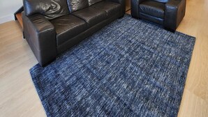 A modern living room with a black leather sofa, a matching armchair, and a blue textured rug. A staircase with a metal railing leads to an upper level. - Semaphore Blue Apartments (Semaphore)