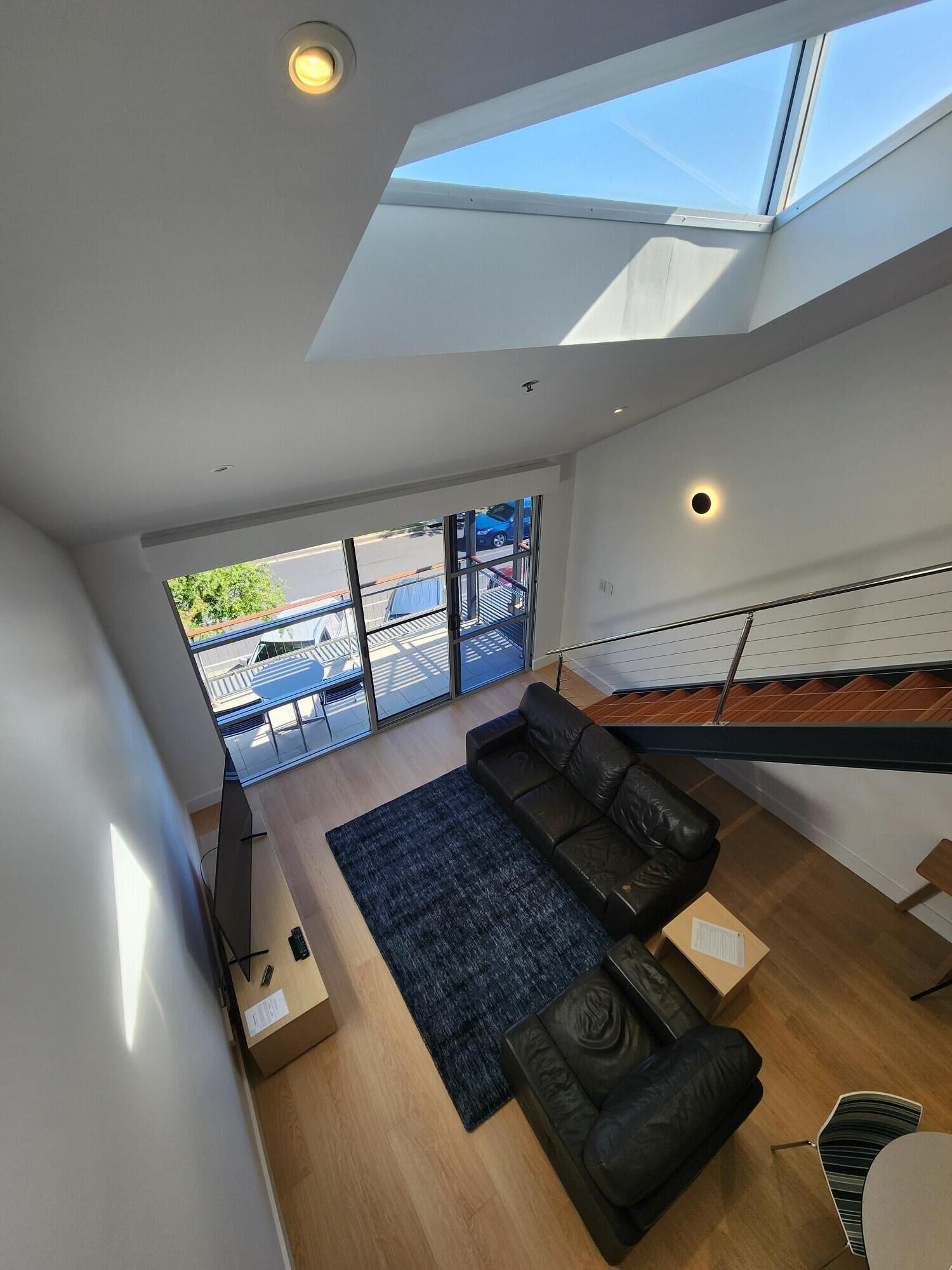 A modern living room with a black leather sofa, a wooden coffee table, and a blue rug. There is a skylight above the sofa and a view of the outside through large windows.