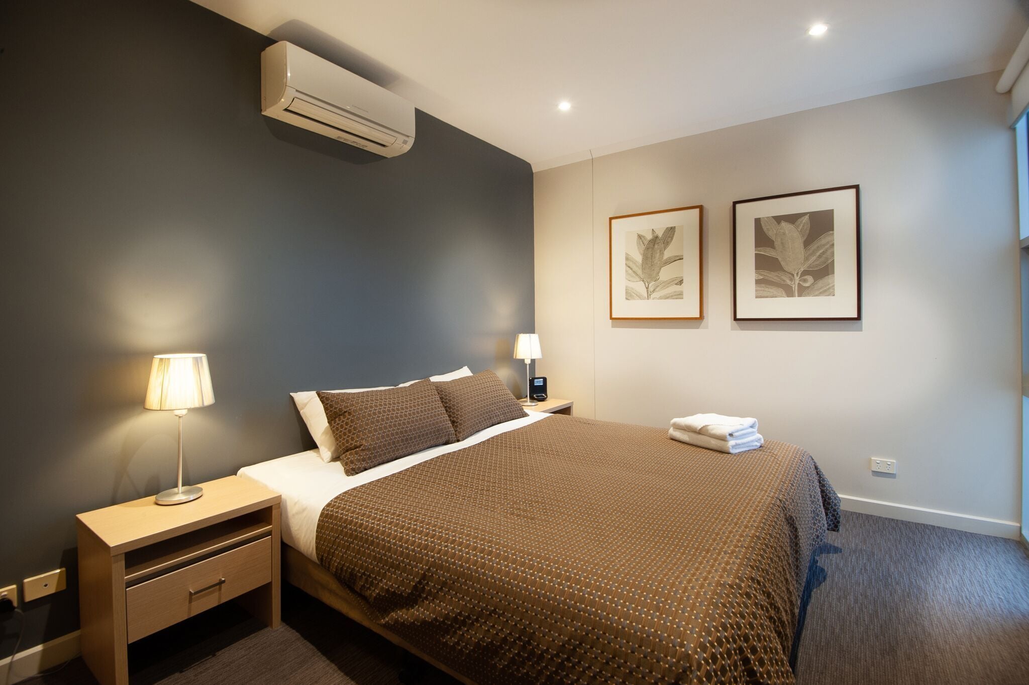 A hotel room with a bed, bedside table, lamp, air conditioning unit, and two framed botanical prints on the wall.