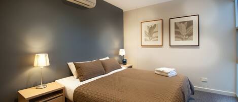 A hotel room with a bed, bedside table, lamp, air conditioning unit, and two framed botanical prints on the wall.