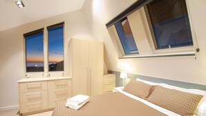 A bedroom with a large bed, a wardrobe, a desk, and two windows with a view of the sunset. - Semaphore Blue Apartments (Semaphore)