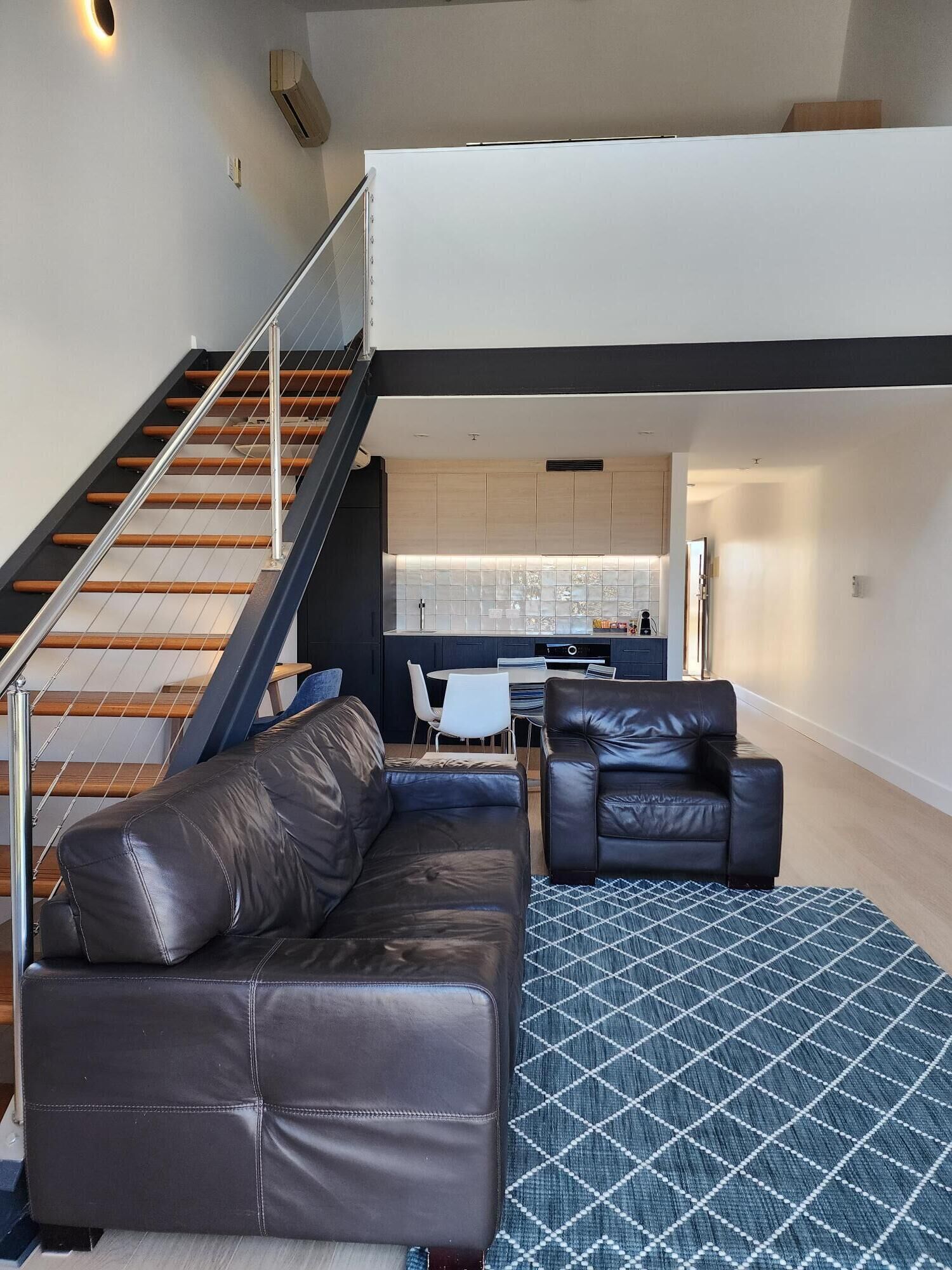 A modern loft apartment with a staircase leading to an upper level, a leather sofa, a patterned rug, and a kitchen area.