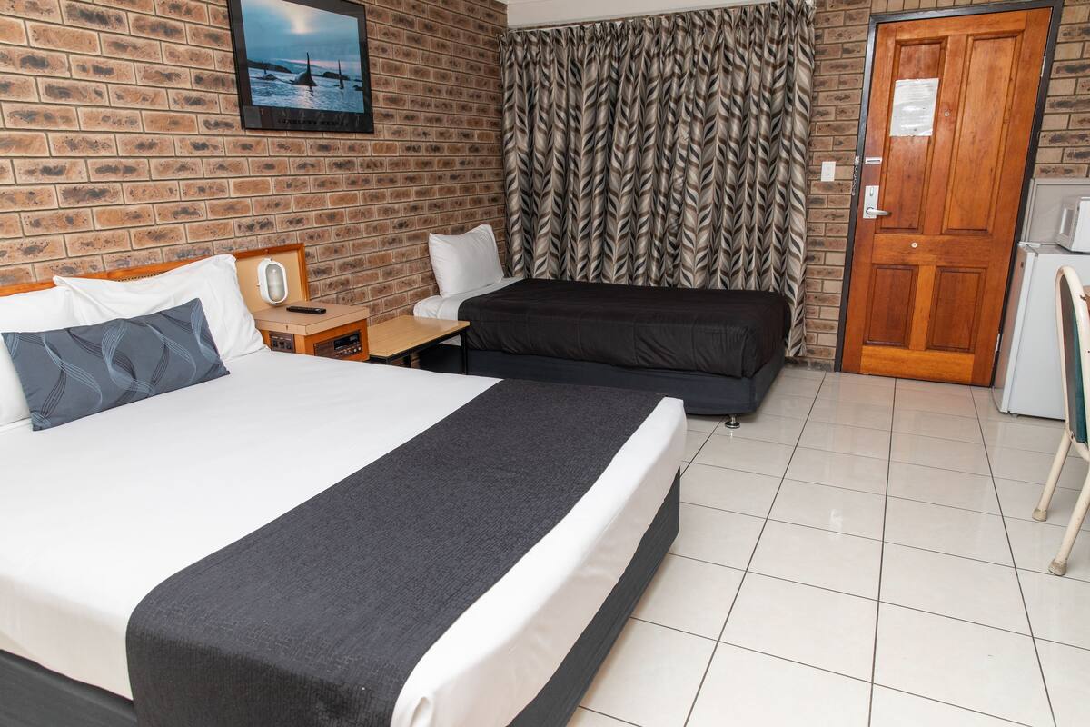 A hotel room with a large bed, a smaller bed, a desk with a chair, a refrigerator, and a brick wall with a picture.