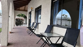 A covered outdoor seating area with tables and chairs, a brick floor, and a view of a fence and trees.