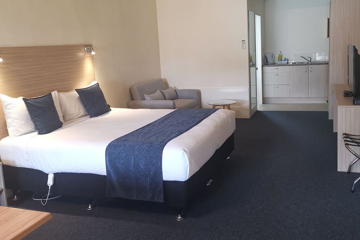 Habitación de hotel con una cama grande, una mesita de noche, una silla, una pequeña mesa, un escritorio, un televisor, una kitchenette y un aire acondicionado.