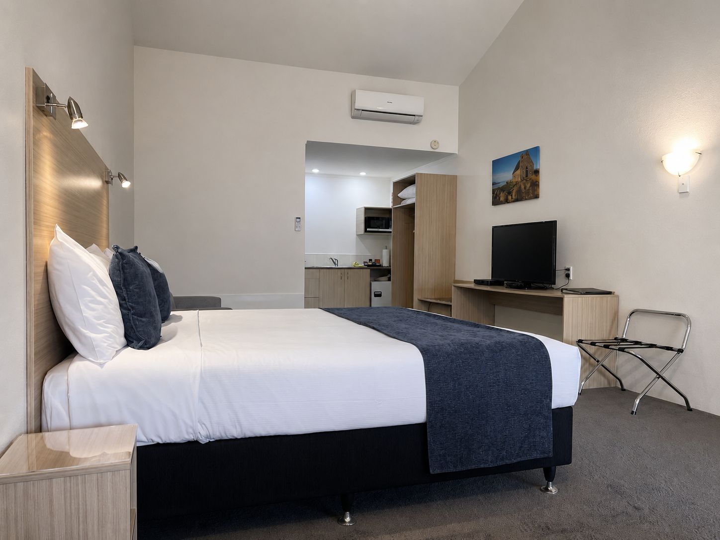 Habitación de hotel con cama, mesitas de noche, un escritorio con televisor de pantalla plana, una silla y una kitchenette al fondo.