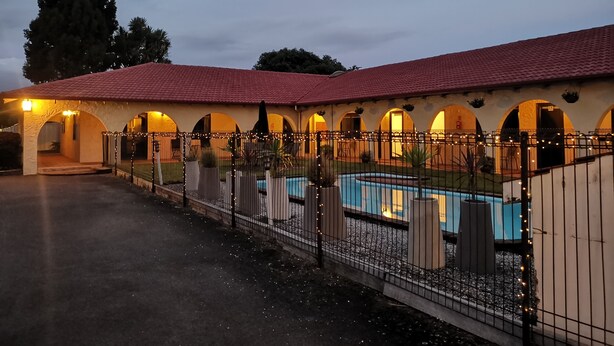 A building with a red-tiled roof, multiple arches, a swimming pool, and a fenced area with string lights. - Captains Quarters Motor Inn (Cambridge)