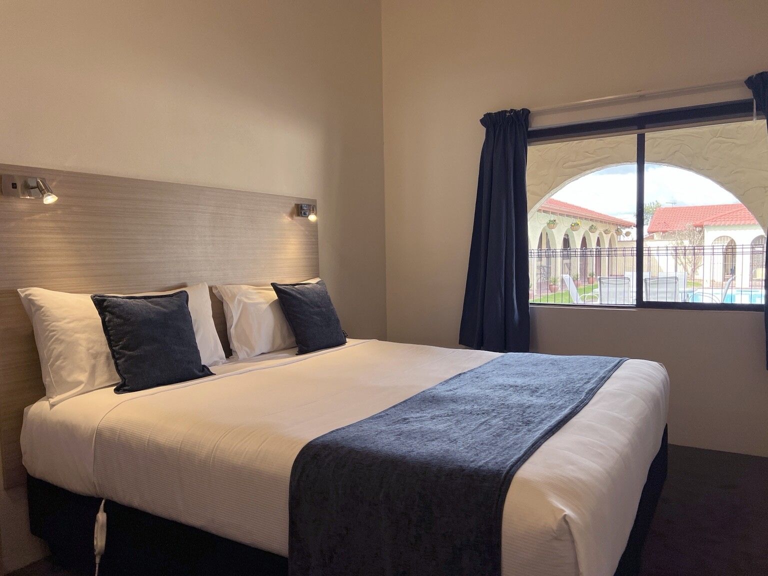 A hotel room with a large bed, a headboard, a window with curtains, and a view of a courtyard.