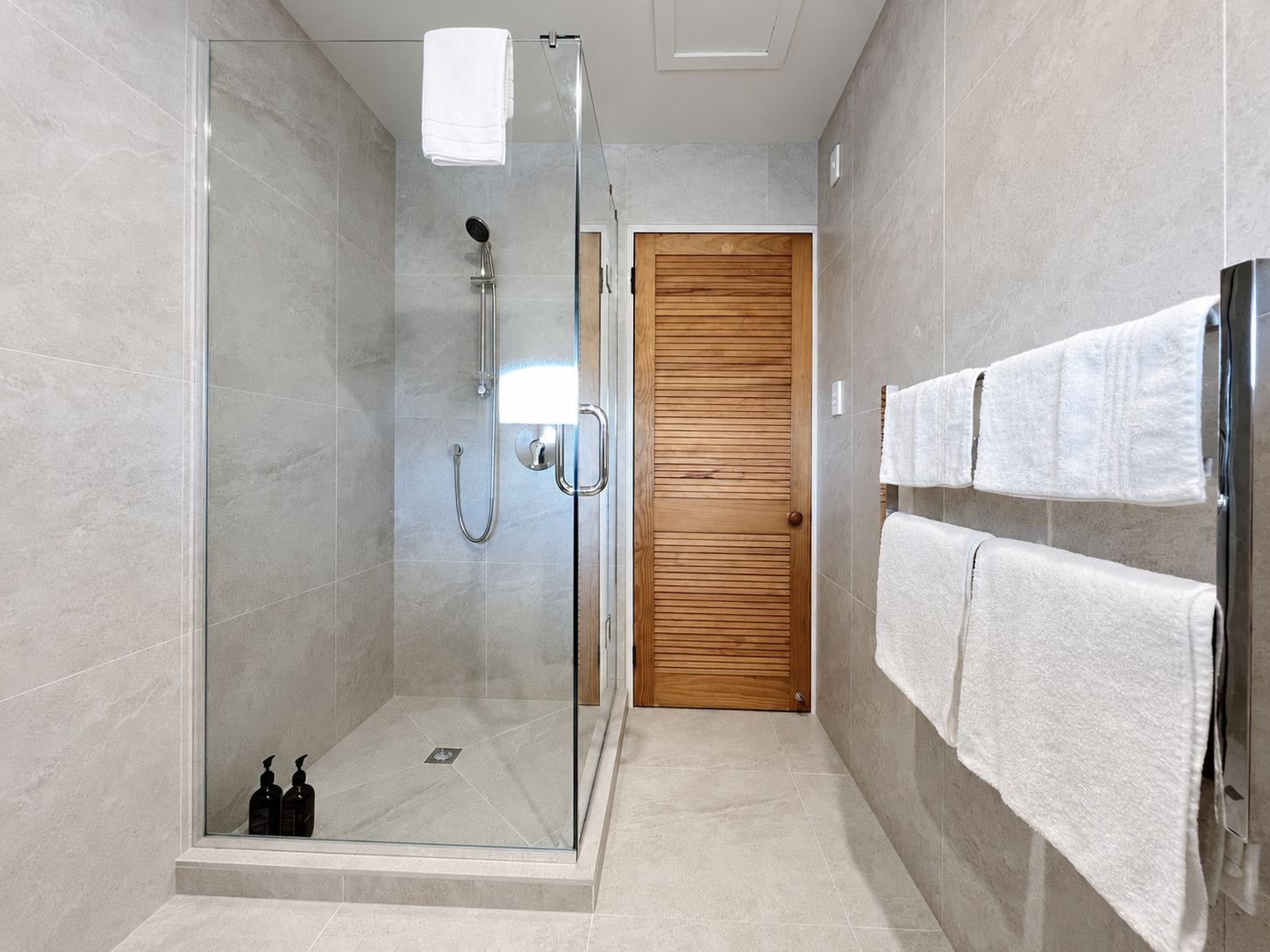 A modern bathroom with a glass-enclosed shower, a wooden door, and a towel rack with multiple towels.