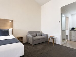 Una habitación de hotel con cama, sofá gris, una mesita y una kitchenette.