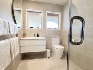 A modern bathroom with a white vanity, two windows, a toilet, and a shower area.