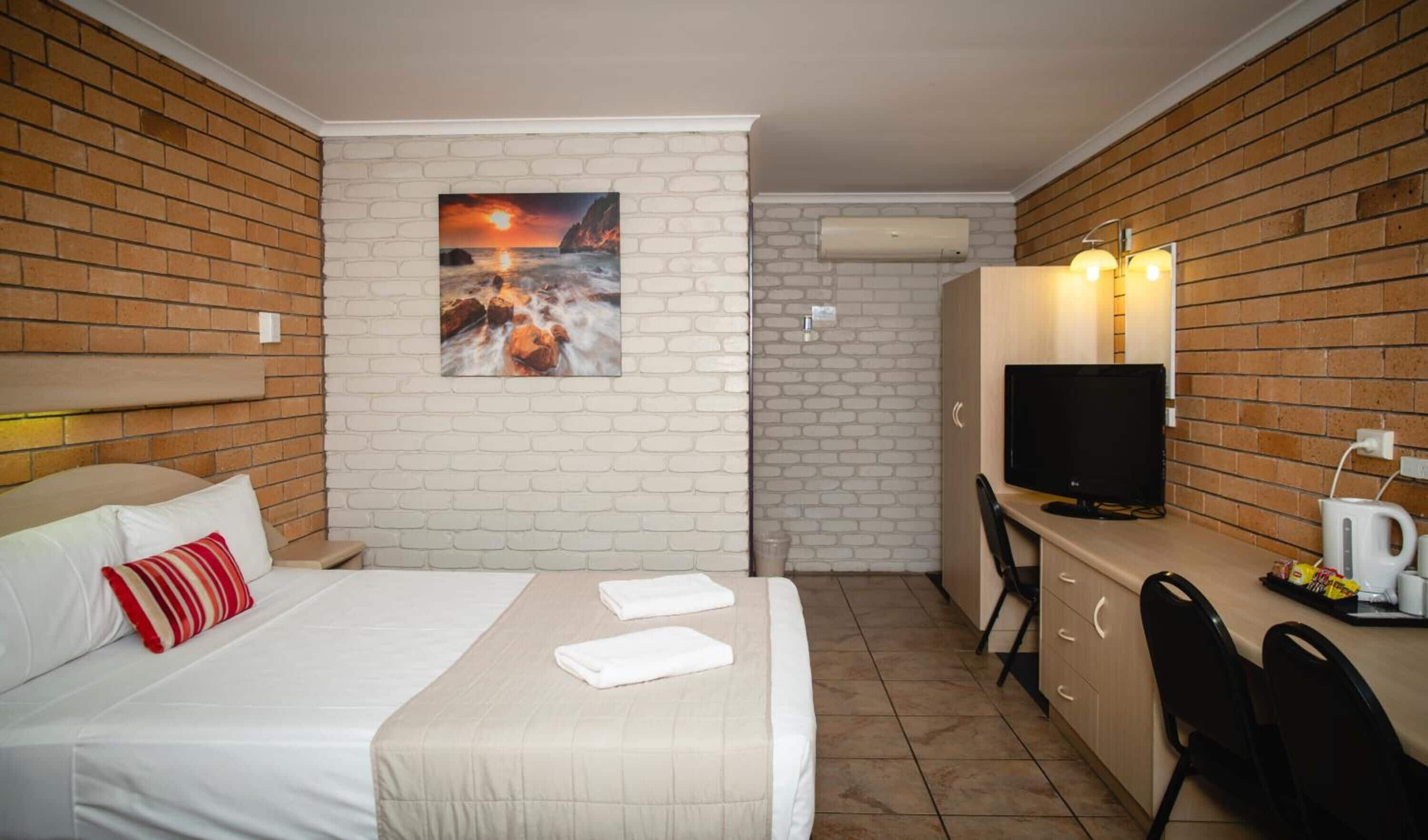 A hotel room with a bed, a desk with a TV, a coffee maker, and a brick wall.