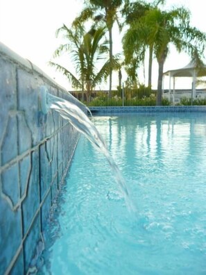 A swimming pool with a water feature, surrounded by palm trees and a fence.