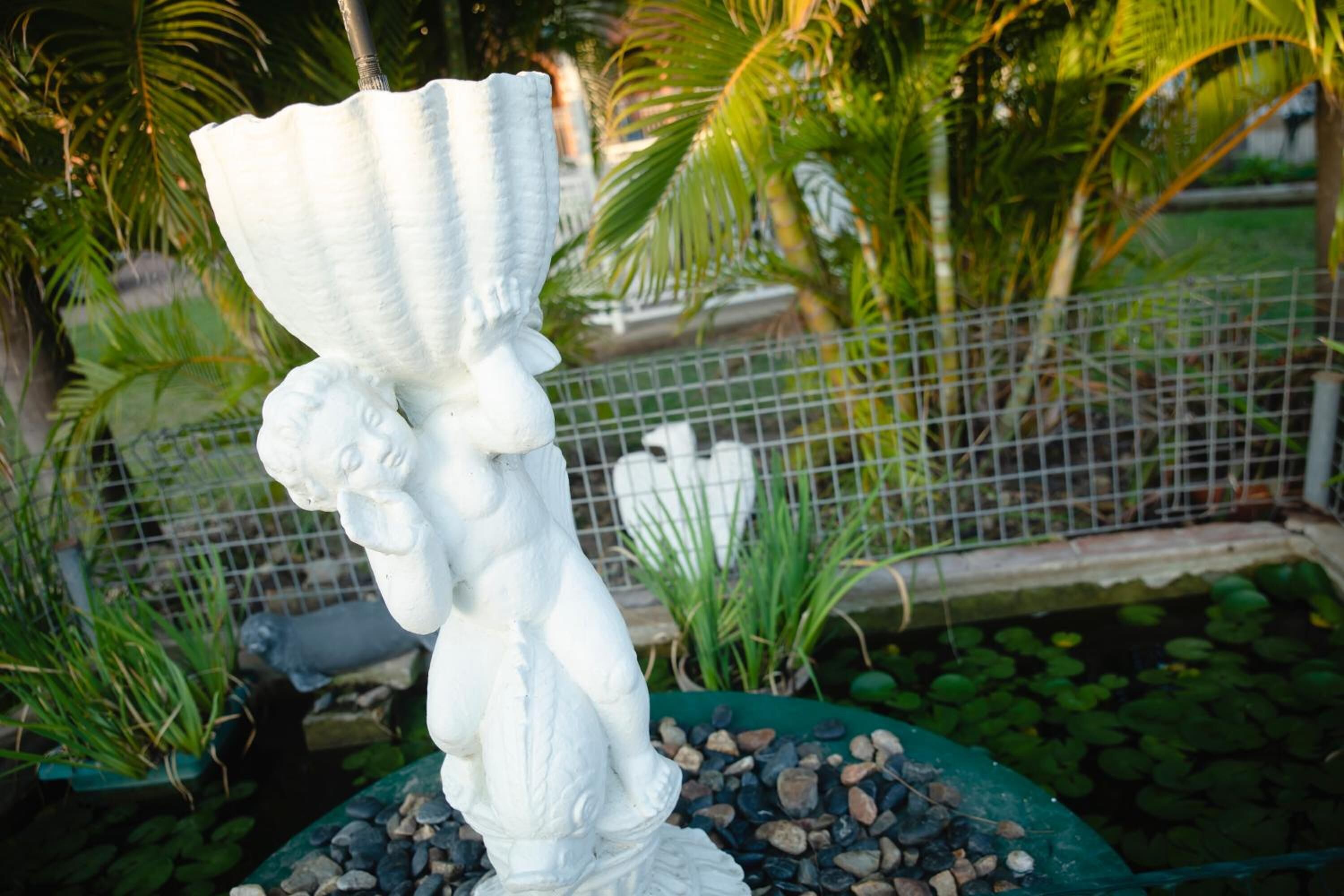Una estatua blanca de una figura sosteniendo un rollo, ubicada sobre un pedestal de rocas y rodeada de follaje y una cerca metálica.