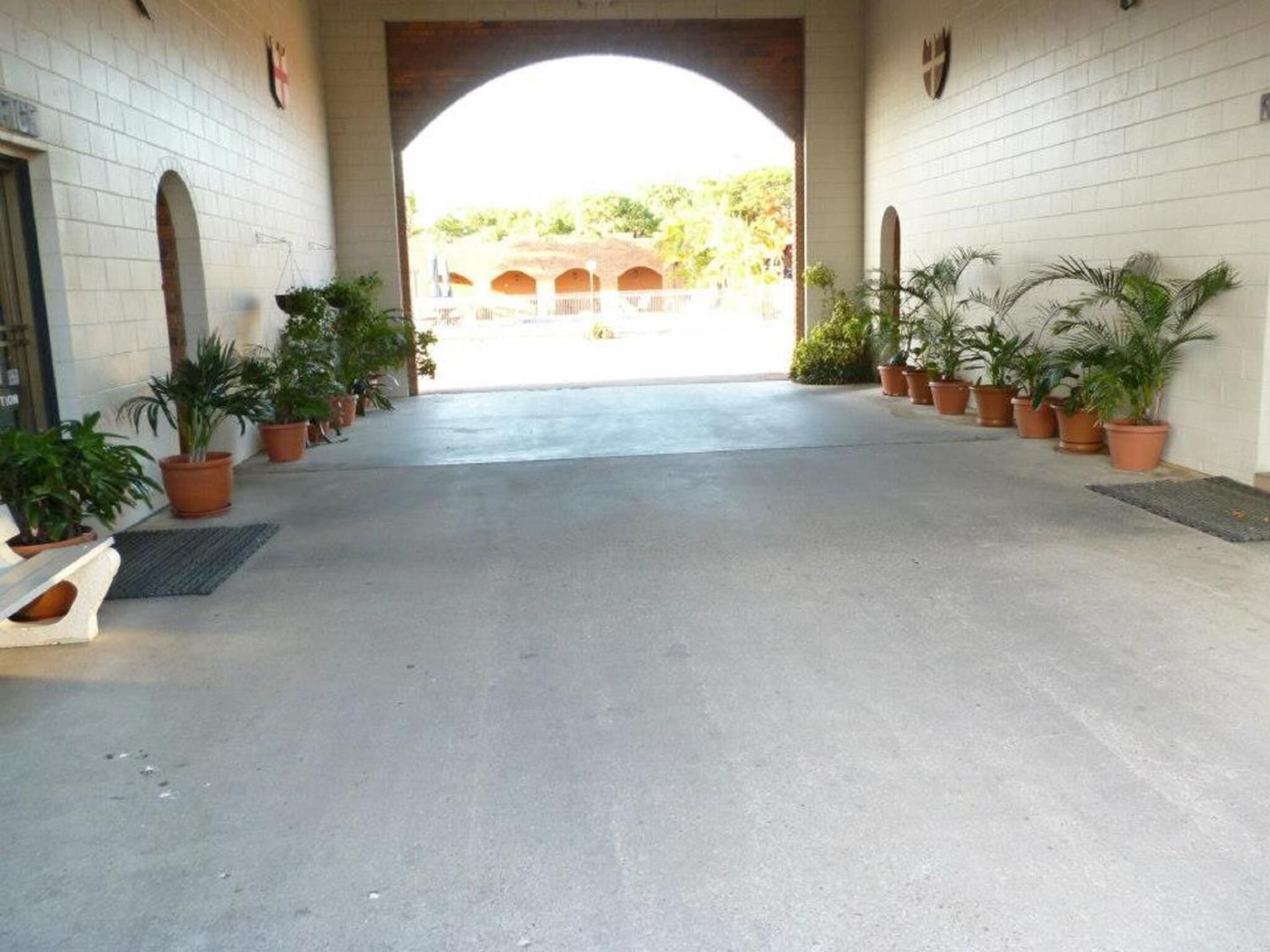 Un pasillo cubierto flanqueado por macetas con plantas, que conduce a un espacio abierto con una estructura de ladrillo al fondo.