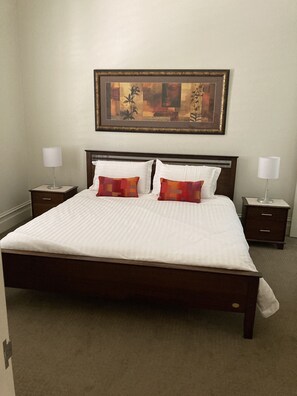 A neatly made bed with white bedding and two red pillows, flanked by two bedside tables with lamps, in a room with a framed artwork above the bed. - Sabina On Little Myers (Geelong)