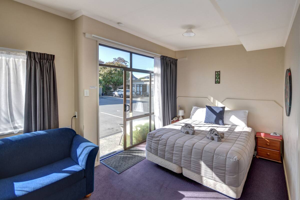 A hotel room with a bed, a blue armchair, a nightstand, and a view of the street through a large window.