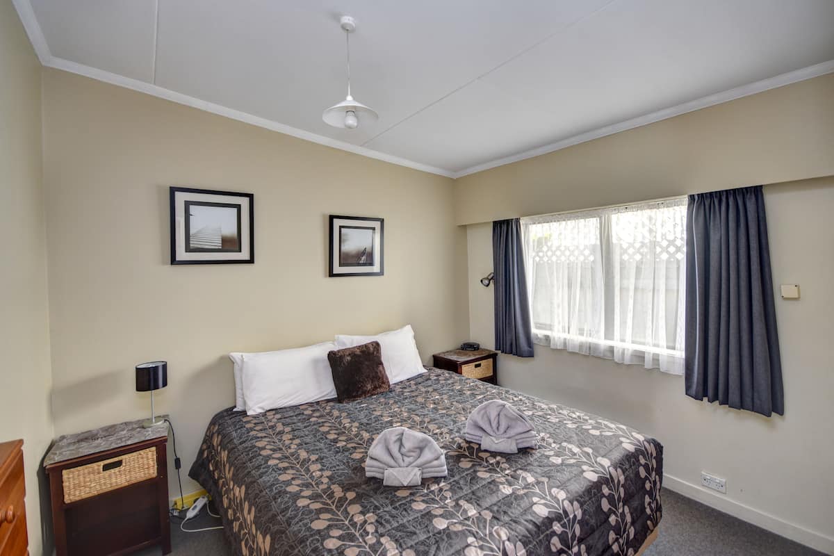 A hotel room with a bed, two framed pictures on the wall, a window with curtains, a bedside lamp, and a small table with a lamp.