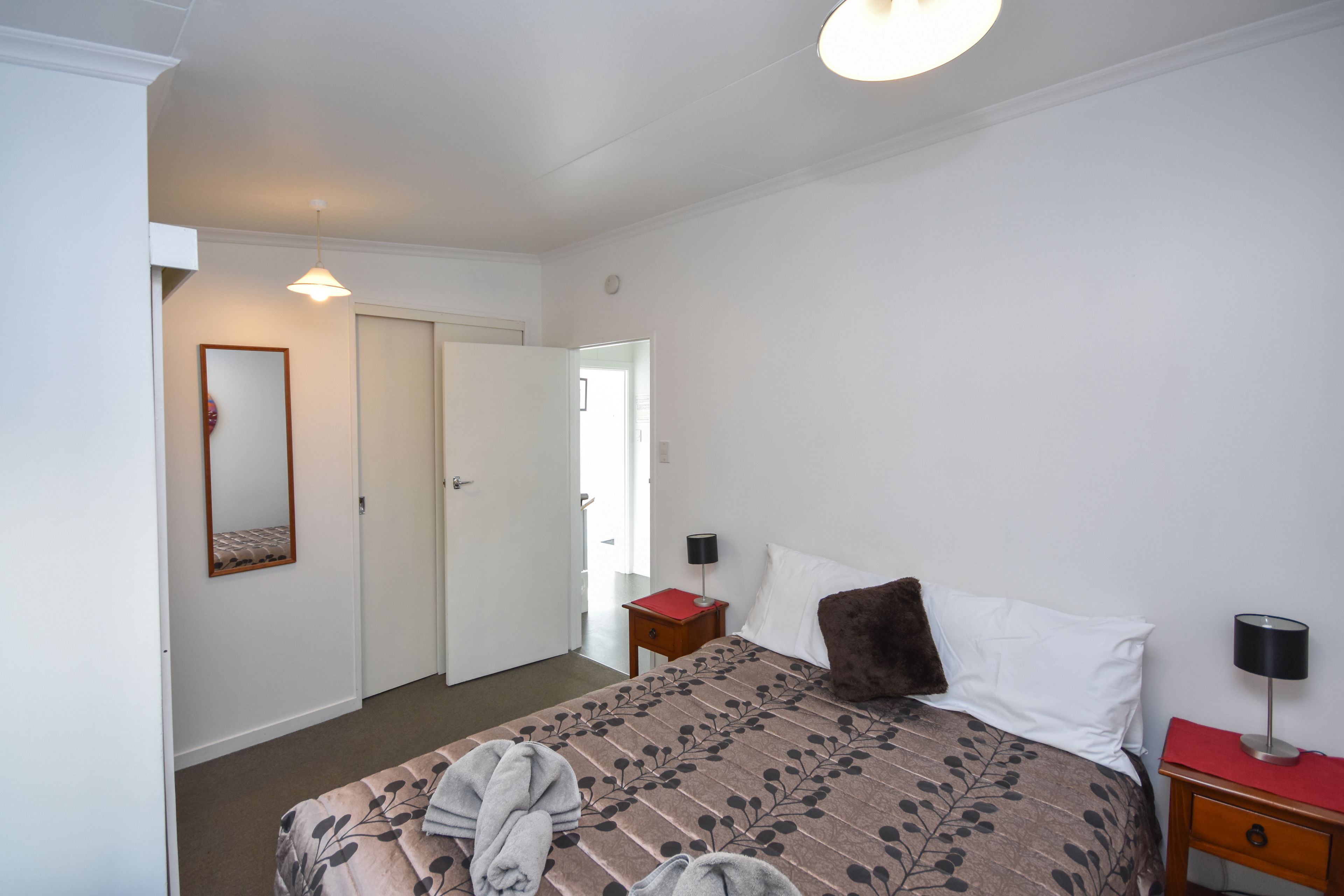 A bedroom with a bed, a bedside table, a nightstand, a lamp, a mirror, and a door.