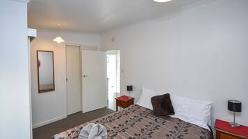 A bedroom with a bed, a bedside table, a nightstand, a lamp, a mirror, and a door.