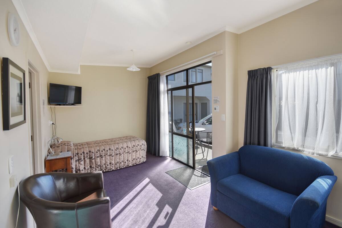 A hotel room with a sofa, a chair, a flat-screen TV mounted on the wall, a window with curtains, and a sliding glass door leading to a balcony.