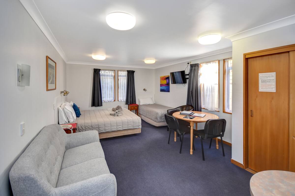 A hotel room with a sofa, a bed, a desk with chairs, a TV, and a window with curtains.