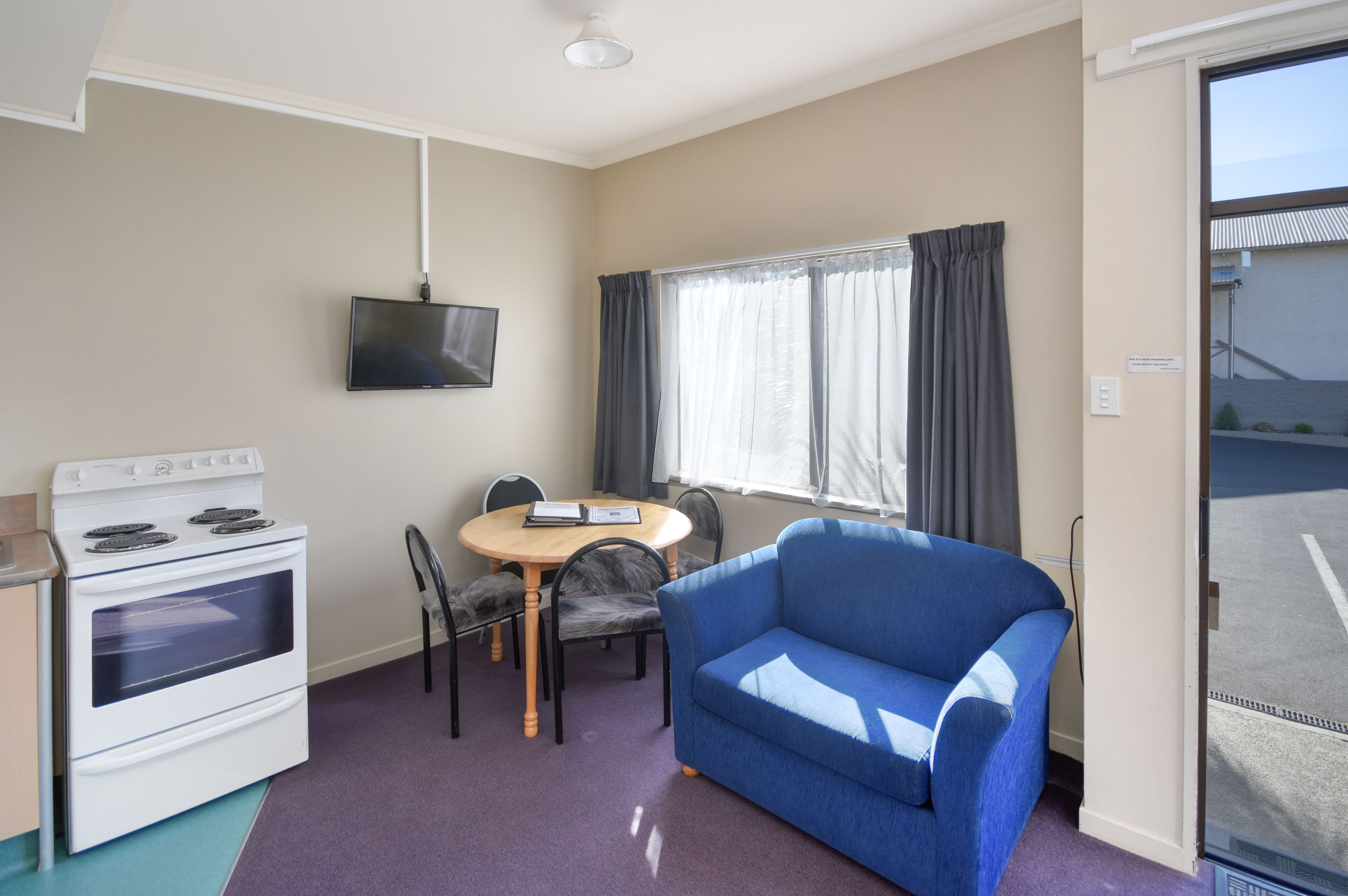 A compact room with a kitchenette, a blue sofa, a dining table with chairs, a TV mounted on the wall, and a window with curtains.