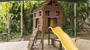 Un’area giochi in legno con scivolo e percorso arrampicata, circondata da una recinzione e vegetazione tropicale.