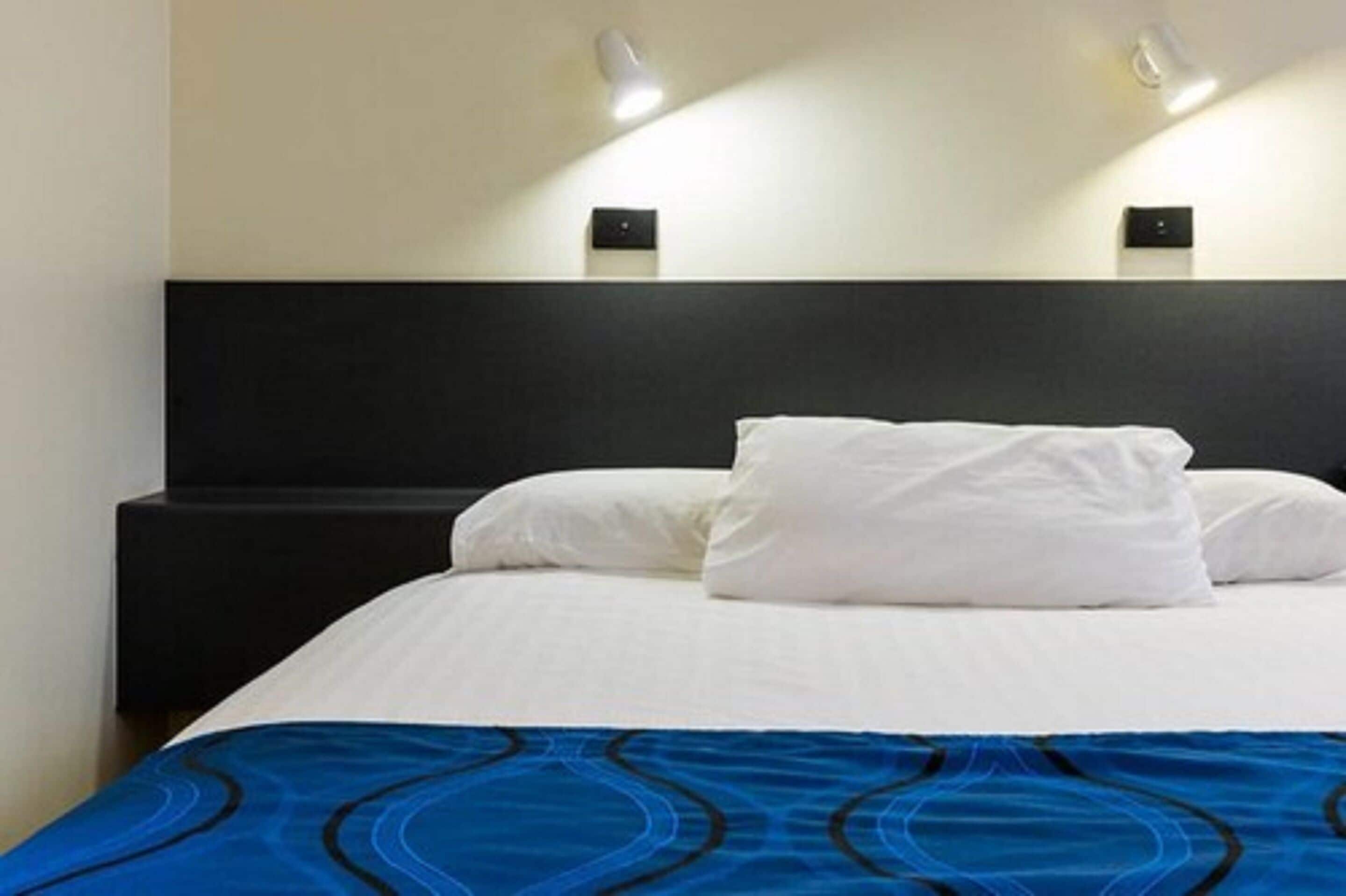 Ein Hotelzimmer mit Bett, weißer Bettwäsche, einem blau gemusterten Überwurf und zwei Wandleuchten.