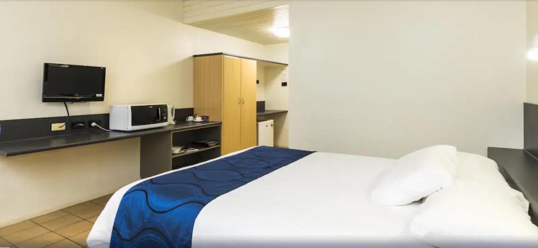 A hotel room with a bed, a television mounted on the wall, a microwave, and a wooden wardrobe.