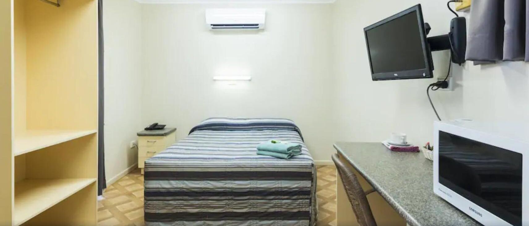 Hotellihuone, jossa on sänky, työpöytä tietokoneella, televisio ja ikkuna, jossa on ilmastointilaite.