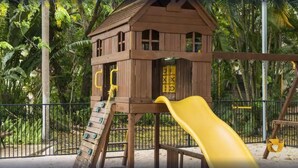 Casetta in legno con scivolo e area giochi, circondata da una recinzione e vegetazione.