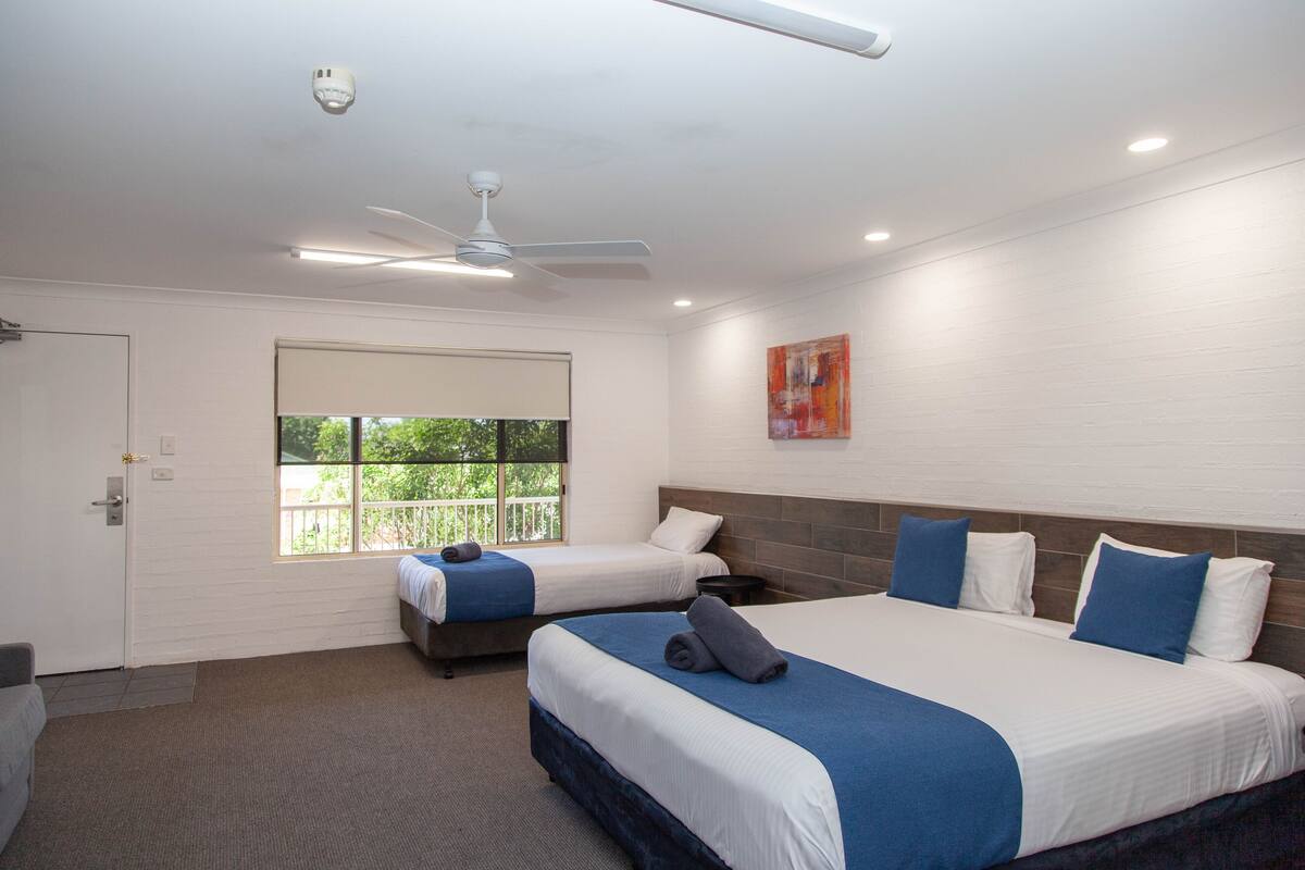 A hotel room with two beds, a ceiling fan, a window with a view, and a painting on the wall.