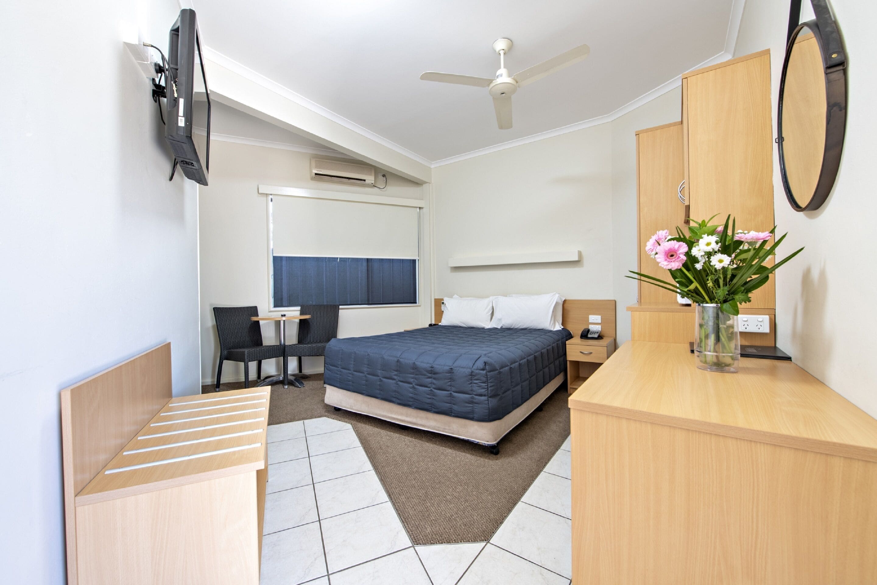 A hotel room with a bed, a desk, a chair, a TV, a ceiling fan, a mirror, and a vase with flowers.