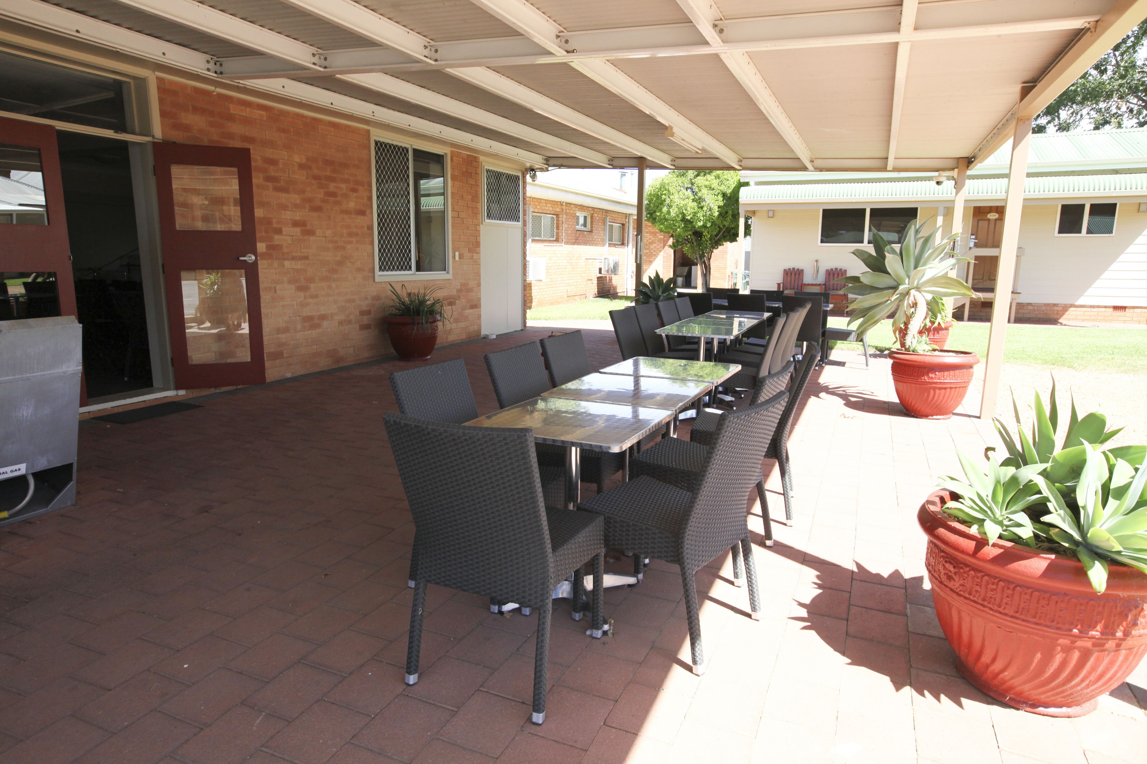 Un patio exterior con una mesa larga y sillas, plantas en macetas y una zona cubierta con pared de ladrillo y una puerta.