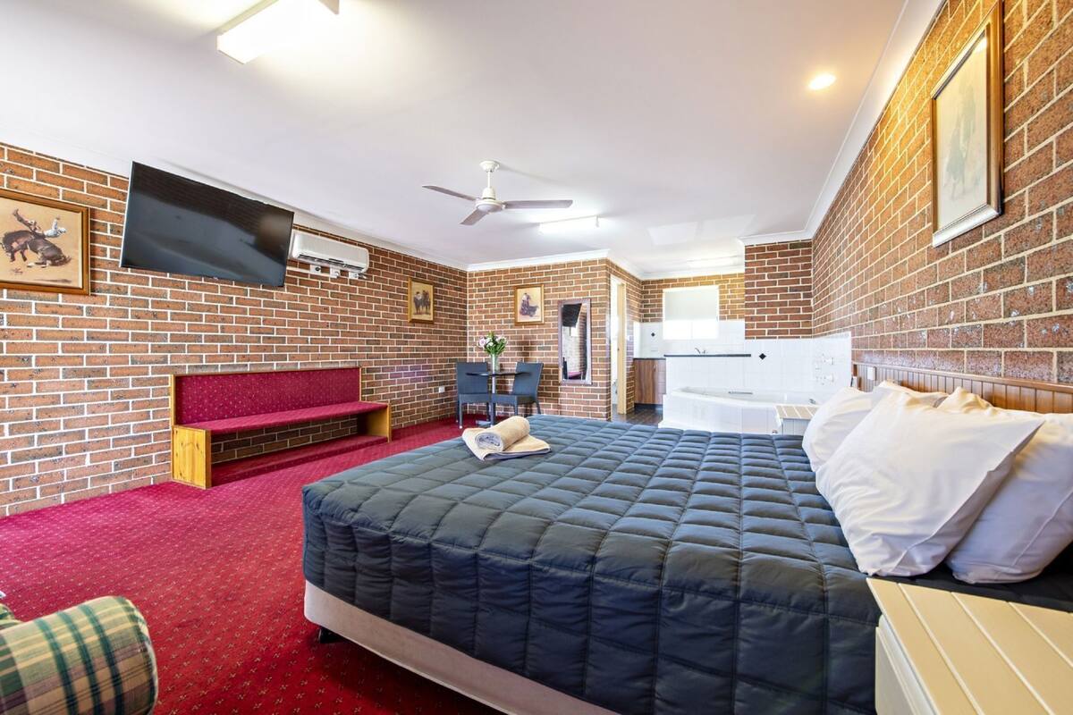 A hotel room with a brick wall, a bed with a quilted mattress, a red carpet, a television mounted on the wall, and a ceiling fan.